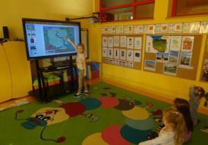 Dziewczynka stoi pod ekranem mobilnym i wskazuje palcem na mapie miasto Warszawę.